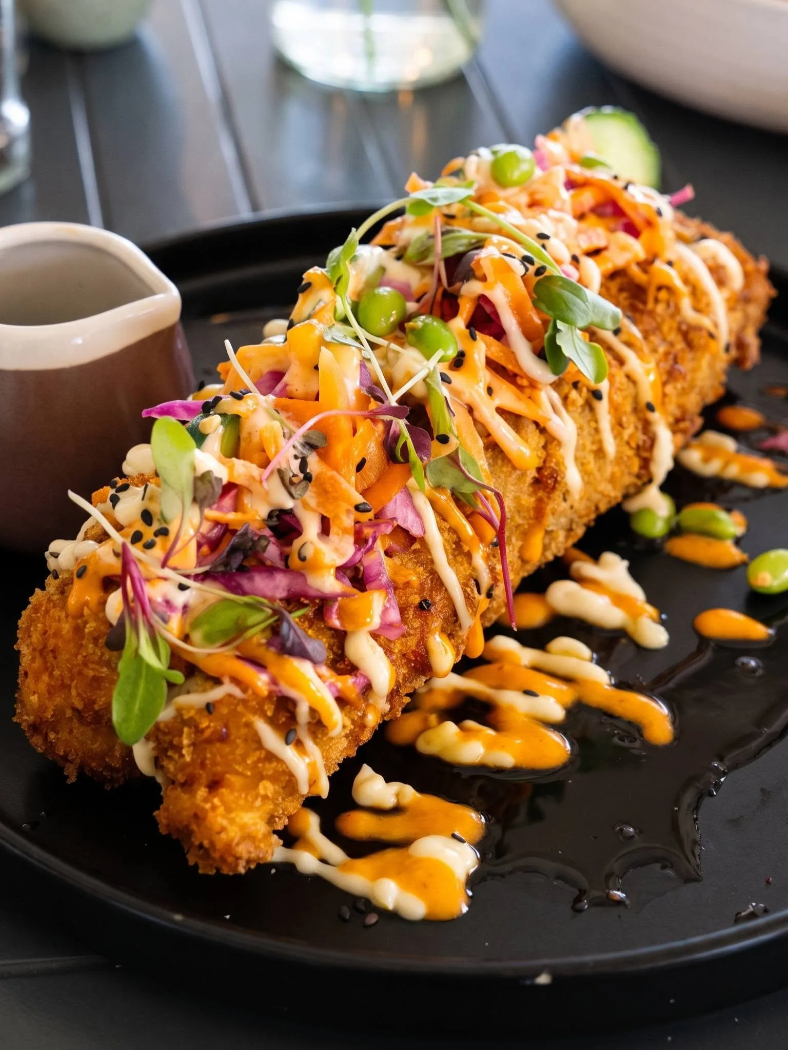 The SushiDogg on a black plate, crumbed and topped with sauces, pickled ginger and greens