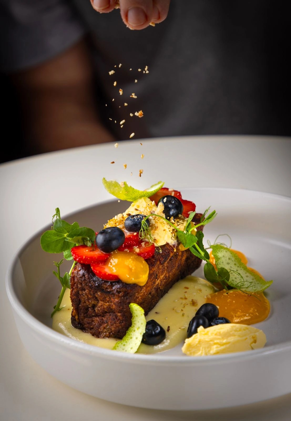 Gold flakes being sprinkled over a plated dessert with berries, mango and cream